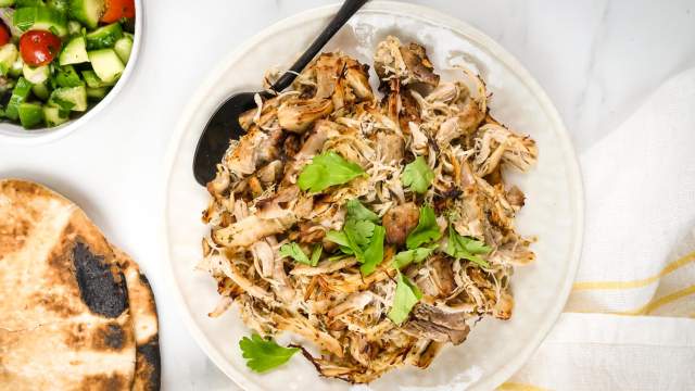 Greek chicken shredded on a plate with oregano, parsley, lemon, pita bread, and cucumber tomato salad.