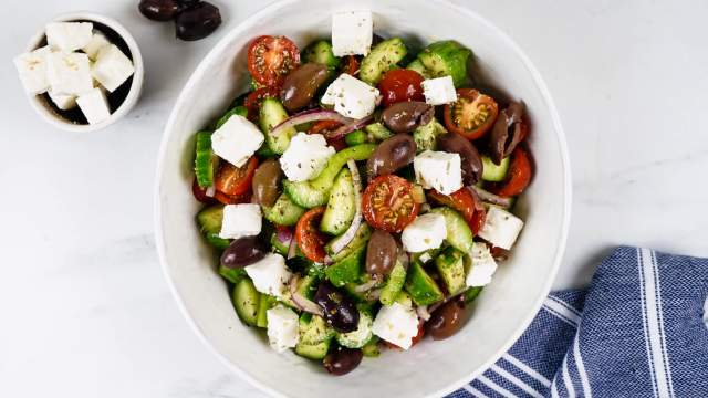 Greek salad with olives, feta cheese, cucumbers, tomatoes, and red onion in a white bowl with a blue napkin. 