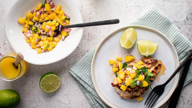 Grilled chicken with mango salsa on a plate with fresh limes and a bowl of mango salsa on the side.