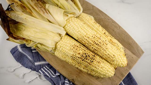 Grilled corn with the husks attached piled on a wood cutting board.