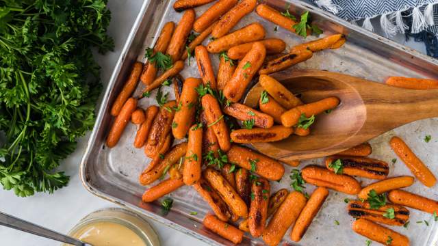 Honey roasted baby carrots with crispy edges on a baking sheet with salt, pepper, honey, and chopped parsley.