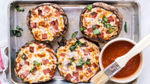 Portobello mushroom pizzas on a baking sheet with marinara sauce, melted cheese, basil, ham, and onions.