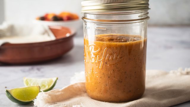 Homemade ranchero sauce in a glass jar with limes and tomatoes on the side.