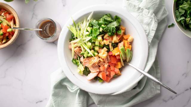 Salmon kale salad with sweet potatoes, avocado, apples, and a maple dijon dressing in a white bowl.