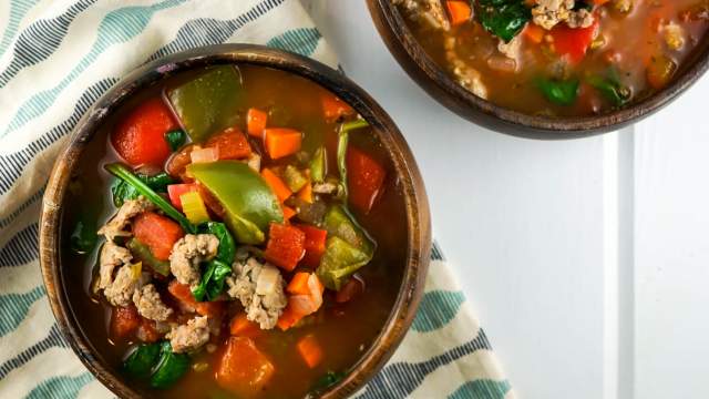 Sausage soup with peppers and spinach in a wooden bowl with carrots, celery, and tomato broth.