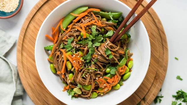 Asian noodle bowls with soba noodles, sesame dressing, carrots, edamame, and snap peas in a white bowl with chopsticks.