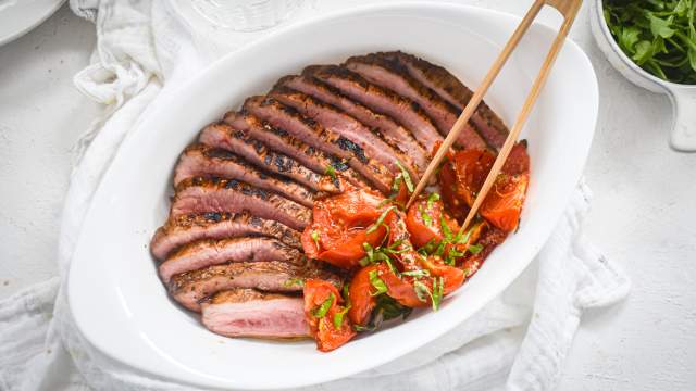 Balsamic flank steak sliced and served with roasted tomatoes in a white plate with wooden tongs.