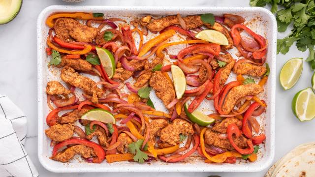 Sheet pan chicken fajitas with chicken breast, peppers, onions, and limes on a baking sheet .