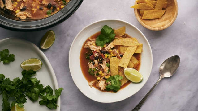 Slow cooker chicken tortilla soup with shredded chicken, beans, corn, cilantro, crispy tortilla strips, and lime wedges.