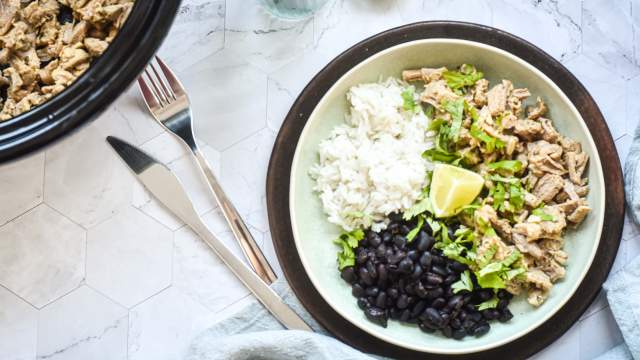 Slow cooker garlic cuban pork shredded on a plate with black beans, white rice, lime, and cilantro.