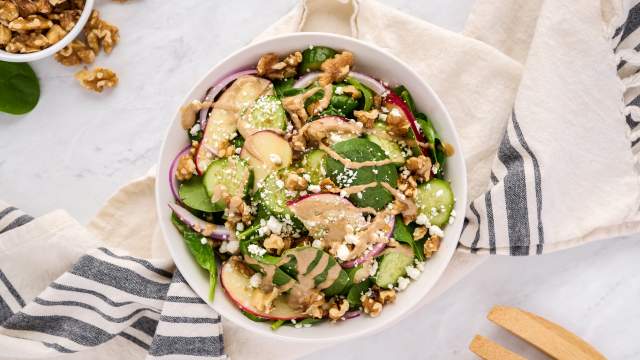 Spinach salad with creamy balsamic vinaigrette, goat cheese, apples, and walnuts in a white bowl with wooden salad tongs.