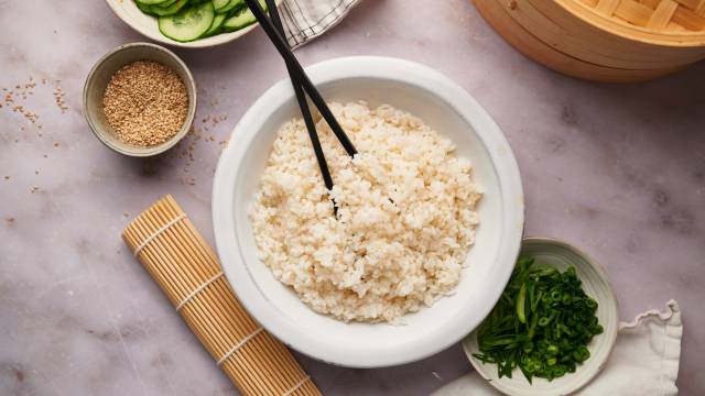 Sushi rice in a bowl with a bamboo mat and sliced vegetables on the side.