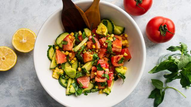 Tomato cucumber salad with corn, avocado, and fresh basil in a bowl with wooden salad tongs.