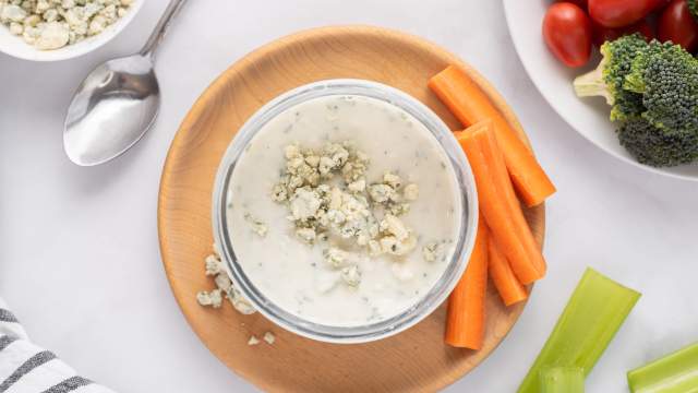 Yogurt blue cheese dressing with crumbled blue cheese in a glass bowl with raw vegetables on the side.